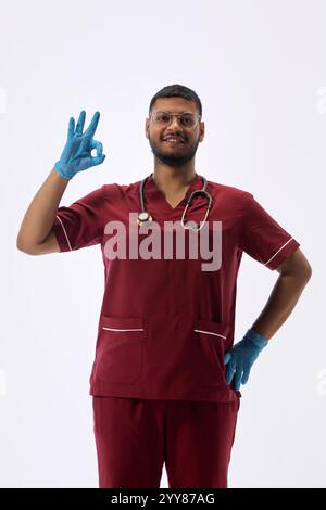 Portrait of young professional, therapist posing confidently holding one hand on hip and gesturing ok sign against white studio background. Stock Photo