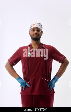 Portrait of of young Indian man, dressed as doctor posing confidently holding hands on hips and looking at camera against white studio background. Stock Photo