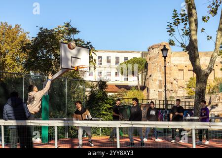 Rome, Italy - Nov 15th, 2024: Boys playing basketball on a court at Parco del Colle Oppio, with the ruins of the Baths of Trajan in the background Stock Photo