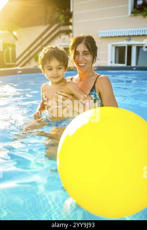 Mother embracing son while playing with yellow inflatable ball in home swimming pool during summer vacation Stock Photo