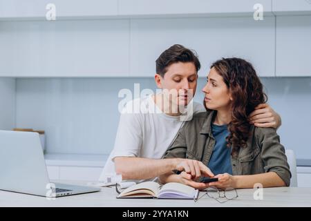 Young couple sitting at kitchen table with laptop, notebook, and calculator, managing home finances and calculating expenses together, concerned about their financial situation Stock Photo