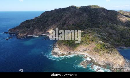 'Aerial drone view of a rugged tropical coastline with lush green hills, rocky shores, and deep blue ocean waters Stock Photo