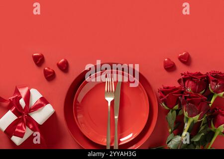Valentine's day. Romantic dinner for date with red plate, chocolate sweets, romantic gift and red roses on red background. View from above. Copy space Stock Photo