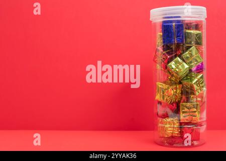 A pile of small, colorful gift boxes wrapped in shiny paper and arranged in a clear container, set against a vibrant red background. Perfect for holid Stock Photo