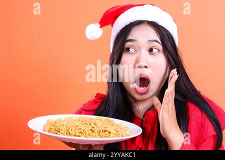 closeup portrait of Asian girl wearing Christmas blouse, Santa hat, surprised to the right, hands holding white plate filled with noodles to the side Stock Photo