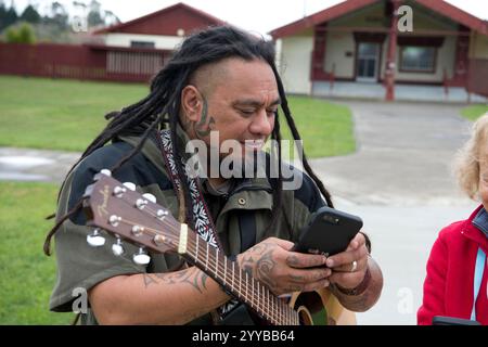 Maori man with guitar using cell phone standing next to a woman wearing a red jacket. Stock Photo