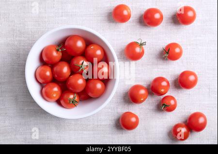 Small red cherry tomatoes in a white bowl on linen fabric, from above. Fresh, ripe, tiny and round cocktail tomatoes of the type Solanum lycopersicum. Stock Photo