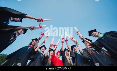Happy students in academic gowns and graduation caps raising their diplomas in celebration of their graduation Stock Photo