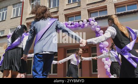 Group of happy students wearing purple ribbons, holding hands and celebrating their last school day in front of their school decorated with balloons Stock Photo