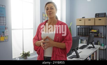 Confident mature woman standing in a modern office, showcasing a professional indoor workplace setting with organized shelves, computers, and office s Stock Photo