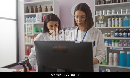 Two hispanic women pharmacists in white coats working together at a pharmacy counter, focusing on the computer screen with various medicines and produ Stock Photo