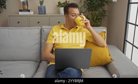 Middle-aged man wearing yellow shirt drinking from a yellow mug while sitting on a couch with a laptop in a cozy living room setting featuring plants Stock Photo