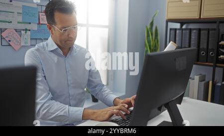 Hispanic man working on a computer in an office environment, focused on his task, with strategy notes and organization charts displayed on the wall. Stock Photo