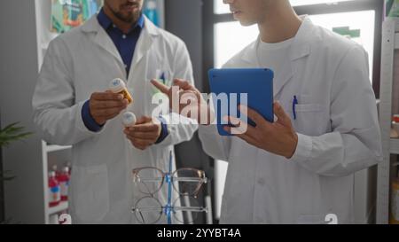 Men pharmacists discussing medication in a pharmacy with one holding bottles and the other using a tablet Stock Photo