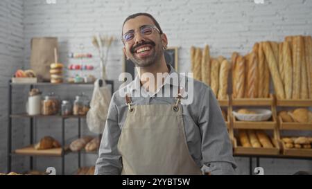 Young man with a moustache smiling in a bakery shop surrounded by various types of bread and pastries displayed on shelves, creating a warm and inviti Stock Photo