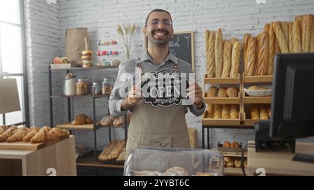 Hispanic man smiling in a bakery holding an open sign with various bread and pastries displayed in the background. Stock Photo