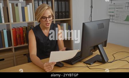 Young woman working in an office with bookshelves behind her, reviewing documents at a desk with a computer Stock Photo