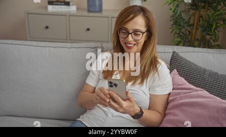 Middle-aged woman sitting on a sofa in a cozy living room, smiling while using her smartphone Stock Photo