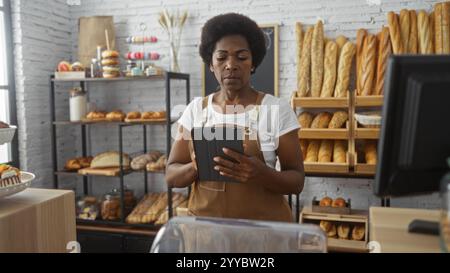 Woman working in a bakery with bread in the background, holding a tablet, showcasing a busy shop environment. Stock Photo