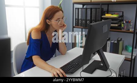 Woman working at a computer in an office, wearing glasses and blue dress, surrounded by shelves and folders, concentrating on her task with a thoughtf Stock Photo