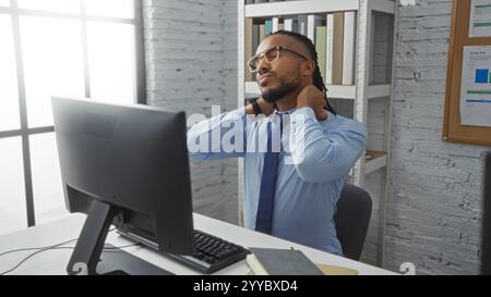 Young man with braids relaxing in modern office workplace, adjusting his neck while seated at a desk with computer monitor in a bright, well-lit room Stock Photo