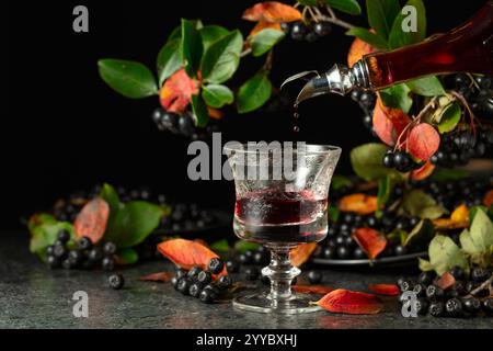 Black chokeberry drink is poured from a bottle into a glass. Sweet drink and fresh berries on a black stone table. Stock Photo