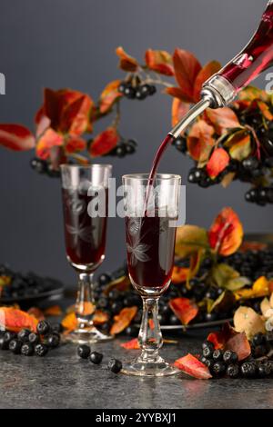 Black chokeberry drink is poured from a bottle into a glass. Sweet drink and fresh berries on a black stone table. Stock Photo