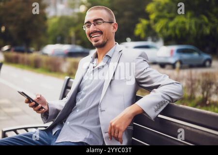 Portrait of cheerful businessman who is messaging on phone. Young businessman sitting on bench in the street and using smartphone. Stock Photo