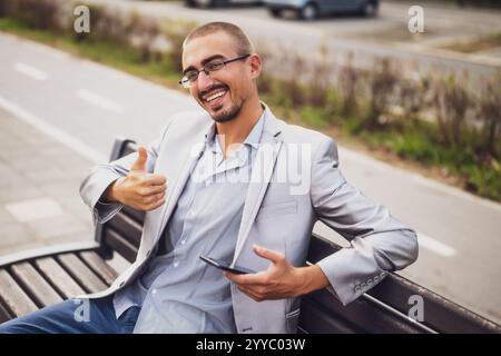 Portrait of cheerful businessman who is messaging on phone. Young businessman sitting on bench in the street and using smartphone. Stock Photo