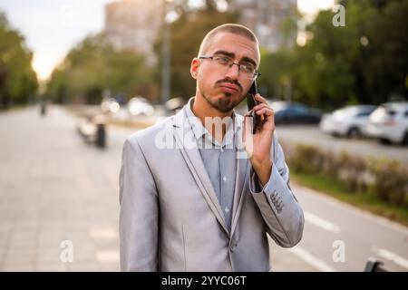 Portrait of distraught businessman who is talking on phone. Young businessman standing in the street and using smartphone. Stock Photo