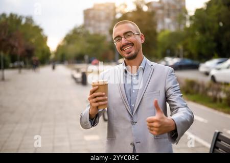 Portrait of cheerful young businessman who is standing in the street, looking at camera and smiling. He is drinking coffee to go. Stock Photo