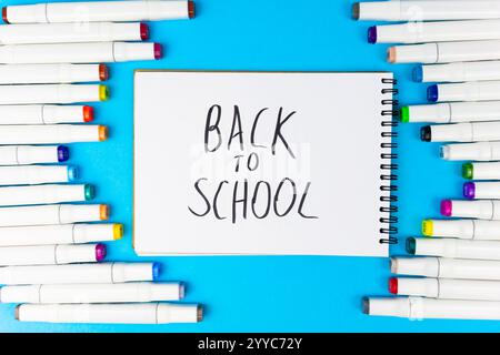 A collection of colorful markers surrounds a spiral notebook. The notebook has the phrase back to school written in bold letters, setting the tone for Stock Photo