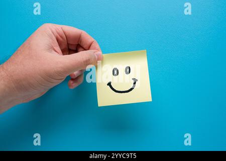 Hand holding yellow sticky note paper with smiley icon on blue background Stock Photo