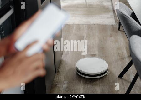 Blurred hands controlling white robot vacuum cleaner via smartphone application while robot vacuum cleaning parquet, focus on robot vacuum cleaner Stock Photo