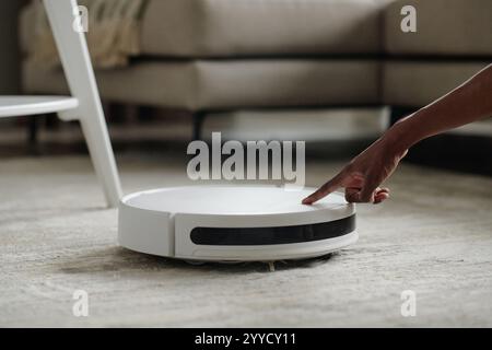 Medium close up of unrecognizable female hand pushing button to turn on white robot vacuum cleaner Stock Photo