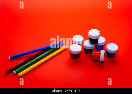A variety of paint pots and markers is arranged on a vibrant red surface. The supplies, including blue, yellow, green, and red colors, invite artistic Stock Photo