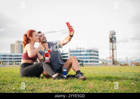 Sporty handocouple sitting on grass taking a selfie with a cellphone after exercising, enjoying outdoor fitness, holding water bottles, and smiling happily, celebrating active lifestyle, health. High quality photo Stock Photo