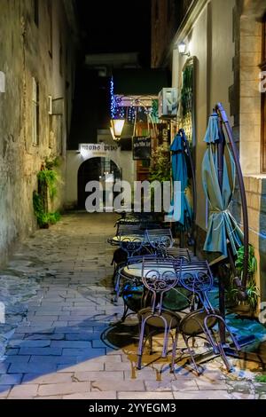 A narrow alleyway with a few tables and chairs outside a restaurant. The tables are empty and the chairs are lined up. The atmosphere is quiet and pea Stock Photo