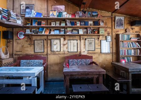 A small cafe with wooden tables and chairs, and a wall full of books. The atmosphere is cozy and inviting Stock Photo