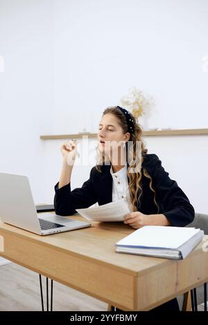 A young woman with long hair thoughtfully examines documents while seated at a modern desk. She appears focused, contemplating ideas. The bright offic Stock Photo