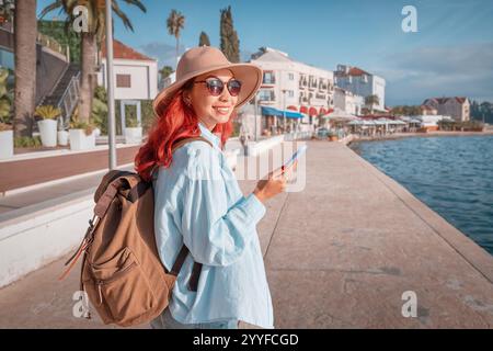 Happy red haired woman using smartphone while walking on seafront promenade near hotels and restaurants Stock Photo
