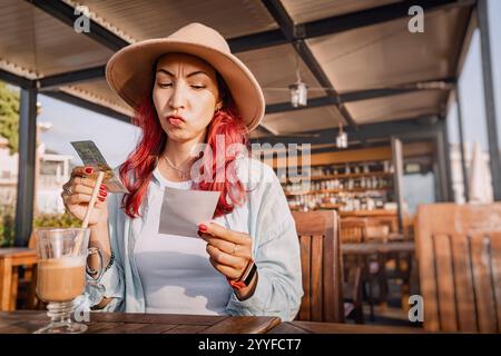 Young woman sitting at a restaurant table with a displeased expression checking the bill and paying in cash Stock Photo