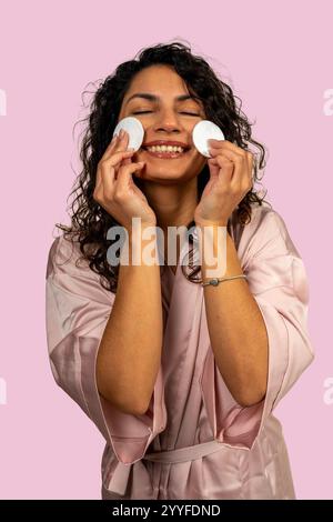 Smiling latin woman wearing bathrobe using cotton pads to remove makeup on pink background Stock Photo