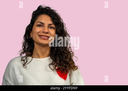 Portrait of a beautiful young woman smiling, wearing a white sweater with a red heart, on a pink background Stock Photo