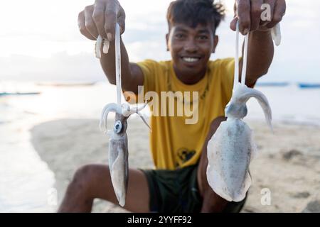 Young Bajau fisherman sitting on sandbank, holding fresh squid he caught, wooden boats in the back. Muna island, Sulawesi, Indonesia Stock Photo
