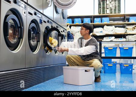 Side view of young Asian man loading clothes into washing machine from laundry basket in self service laundry, household chores concept Stock Photo