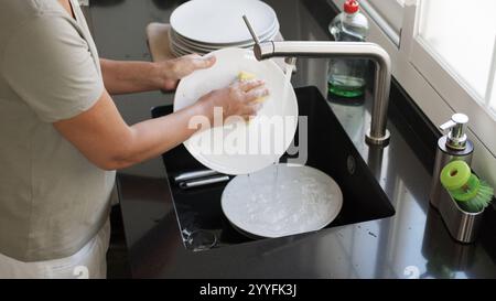 Woman washing and rinsing dishes by hand under a stream of water in the kitchen sink. Washing dishes process. Stock Photo