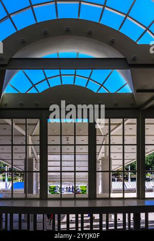 A large building with a glass ceiling and a view of a courtyard. The courtyard is filled with people and a few benches Stock Photo