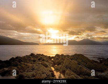 Sunrise over water by a mountain and sea landscape with golden sun beams shining through low clouds, with rocks in the foreground, at Isle of Skye Stock Photo
