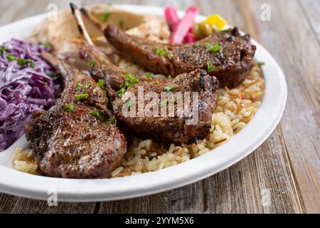 A view of a plate of lamb chops, featuring rice pilaf, cabbage salad, and hummus. Stock Photo